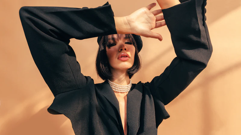 A woman in a black blazer and pearl necklace posing against a tan wall