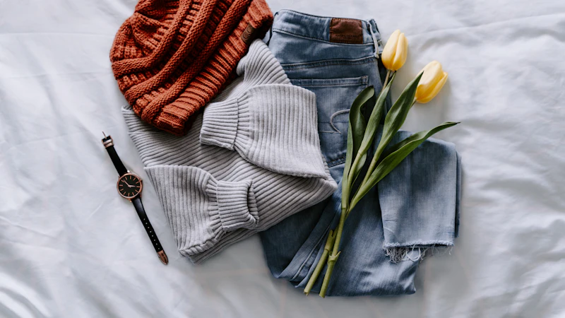 A rust-colored beanie, grey sweater, blue jeans, wristwatch, and yellow tulips on white bedding