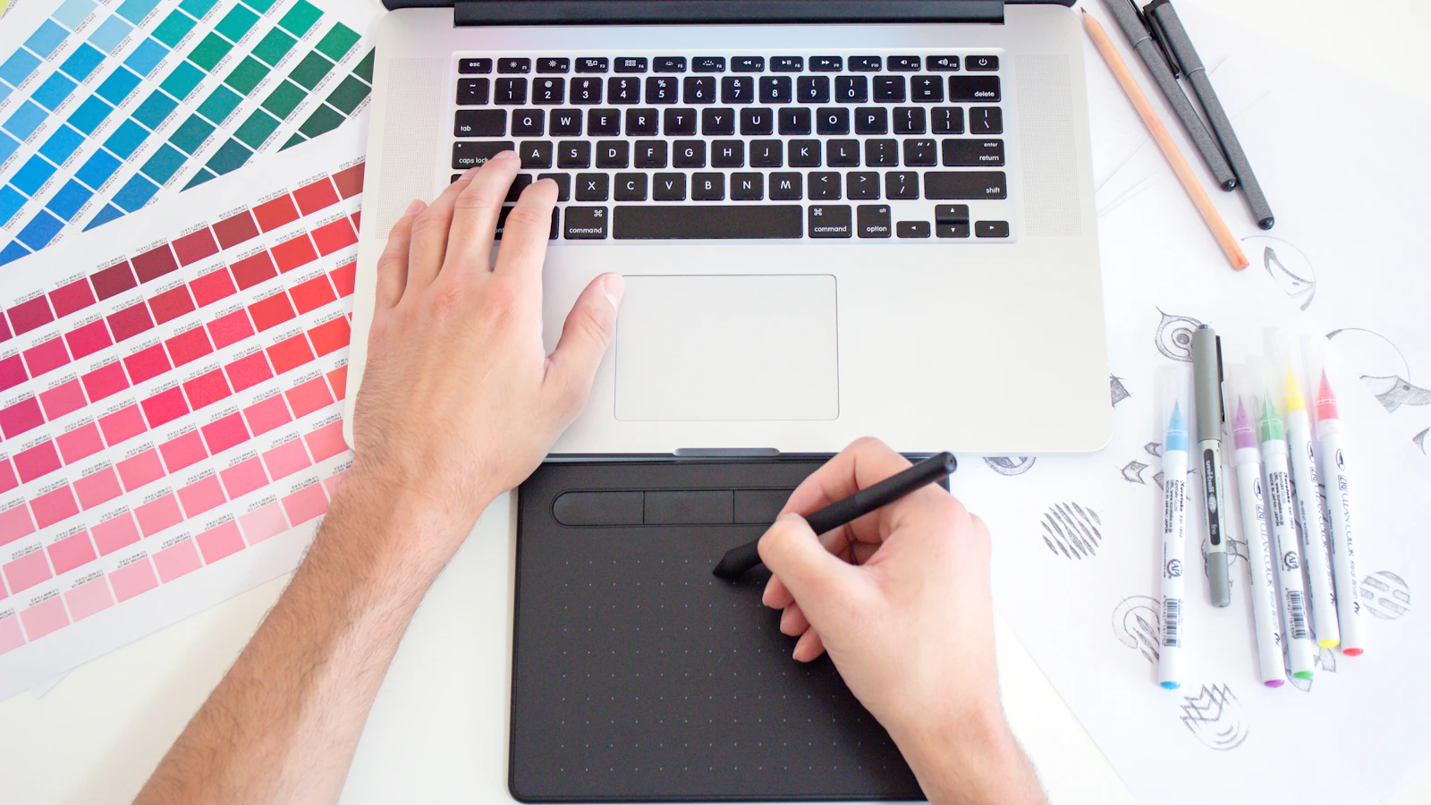 A person using a digital drawing tablet and laptop with color swatches and markers